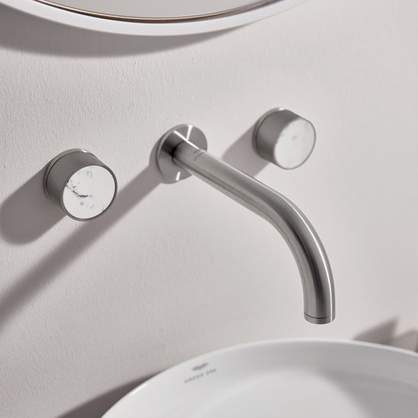 A modern wall-mounted faucet Atrio set with a curved polished chrome spout and two round handles, shown installed above a white sink against a light beige wall.